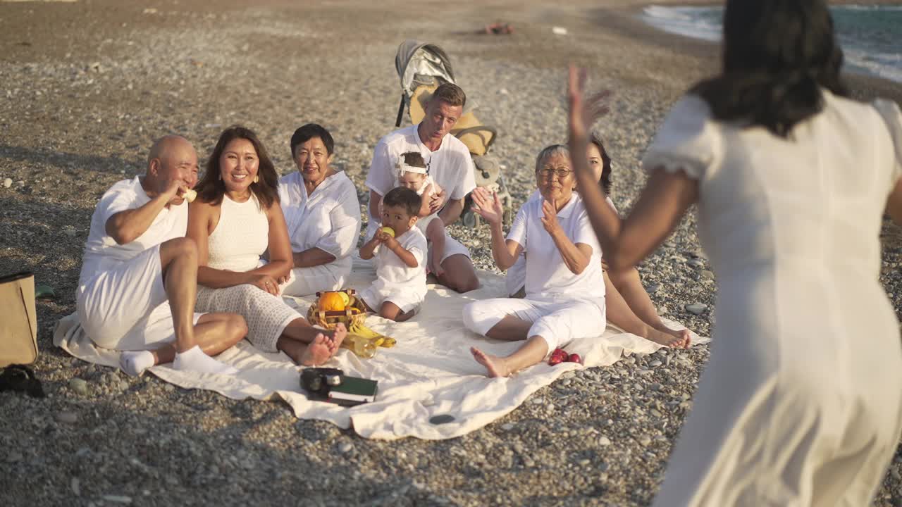 笑得开心的亚裔家庭和白人男子猜词，作为女人手势慢动作。积极的多代人在地中海海滩上享受日落乐趣的广角镜头。视频素材
