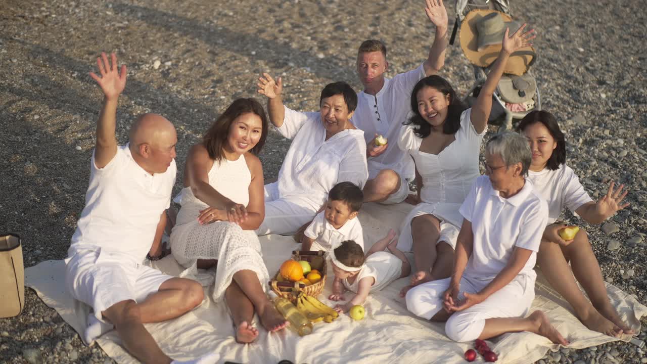 日落时分，坐在地中海海岸的卵石上，欢快的亚裔家庭和白人男子一边挥手一边看着相机。幸福家庭享受假期，户外休闲。视频素材