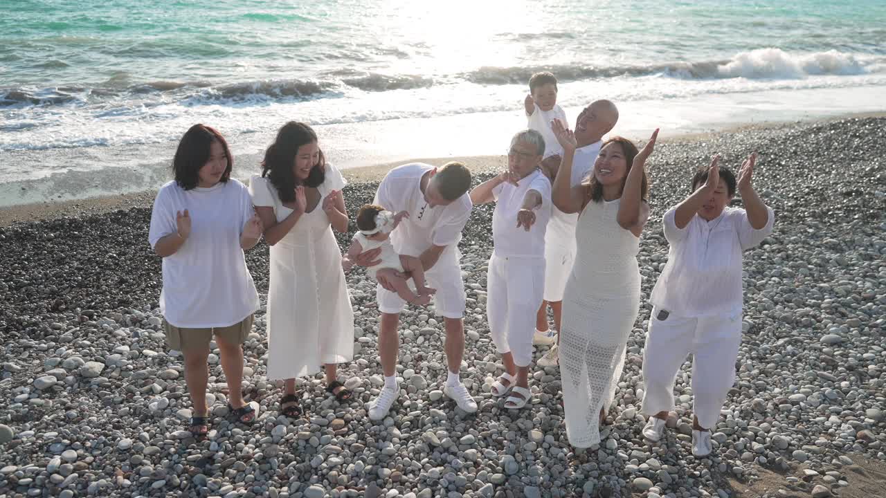 欢快的亚洲家庭拍手，在慢动作中跳舞，与蓝绿色的地中海海浪飞溅在鹅卵石上的背景反射日落的阳光笑。幸福与旅游理念。视频素材