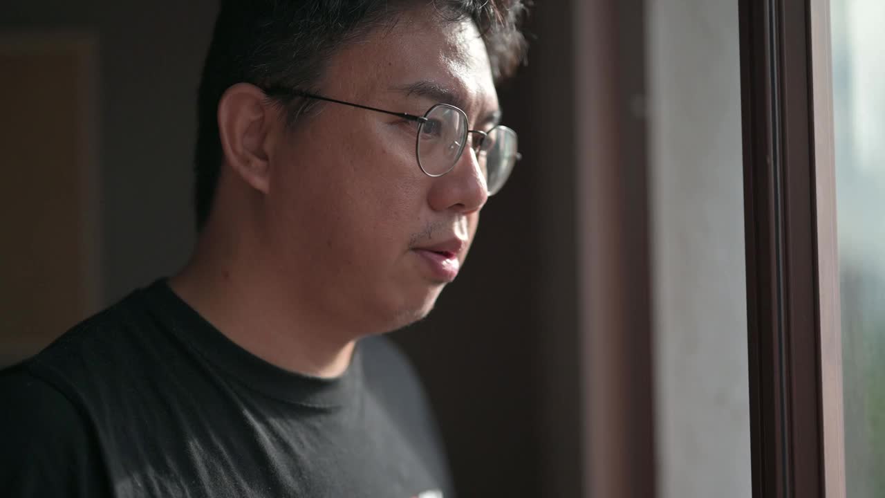 一名亚洲华人中年男子在夕阳西下时从家中望向窗外，脸上带着严肃的表情，手里拿着一个热饮杯视频素材