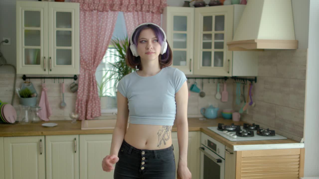 年轻漂亮的女人戴着耳机在家里的厨房跳舞视频下载
