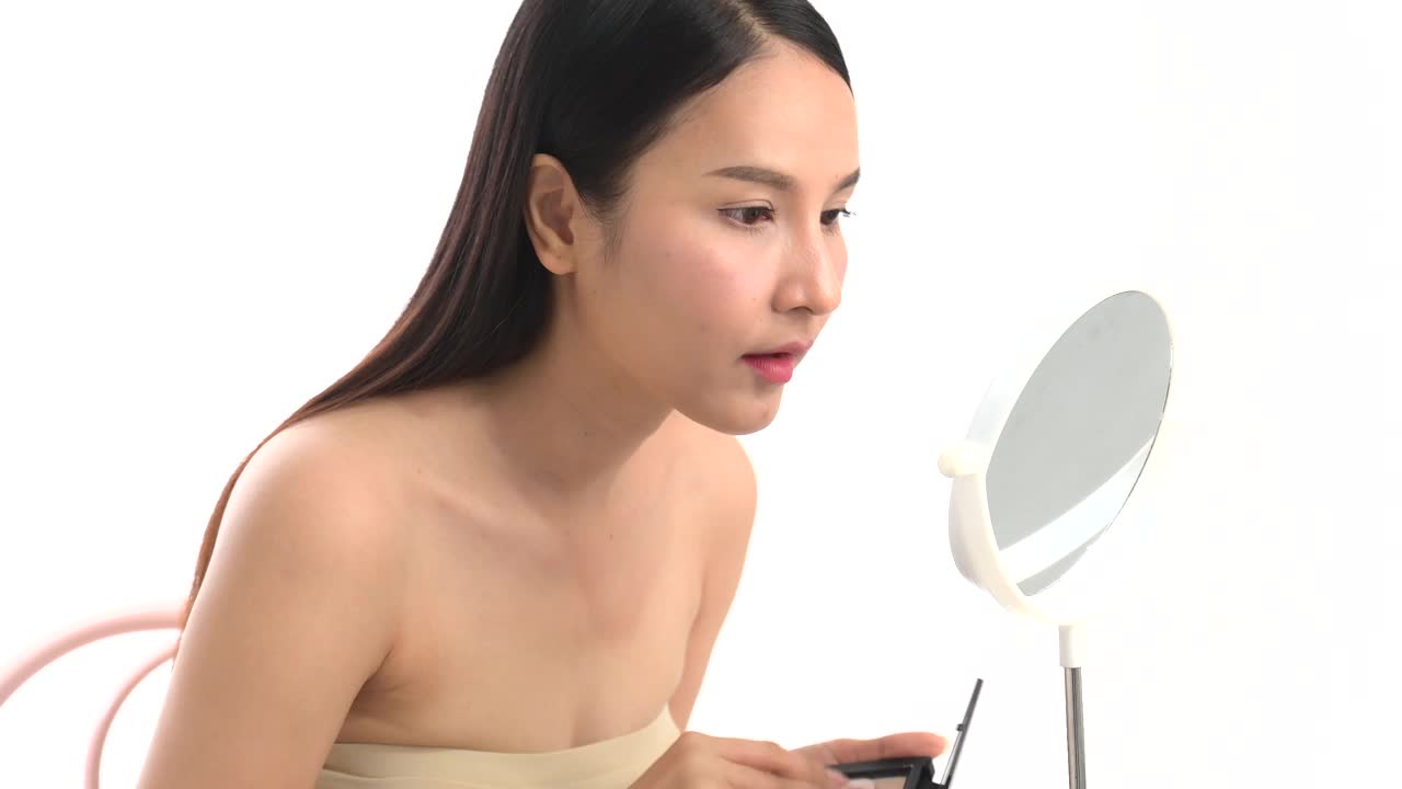 亚洲女性喜欢自己化妆。年轻迷人的美丽感到快乐的看着镜子和着色眉毛在脸上享受美丽的化妆在白色的背景视频购买