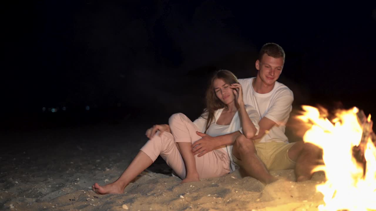 一对年轻的夫妇正在火边休息，坐在沙滩上，互相拥抱，愉快地聊天。视频下载