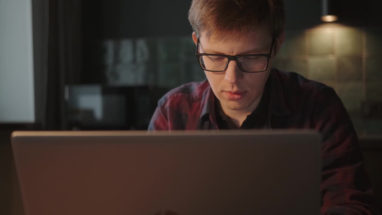 晚上，一名戴眼镜的男子在家里用笔记本电脑工作视频下载