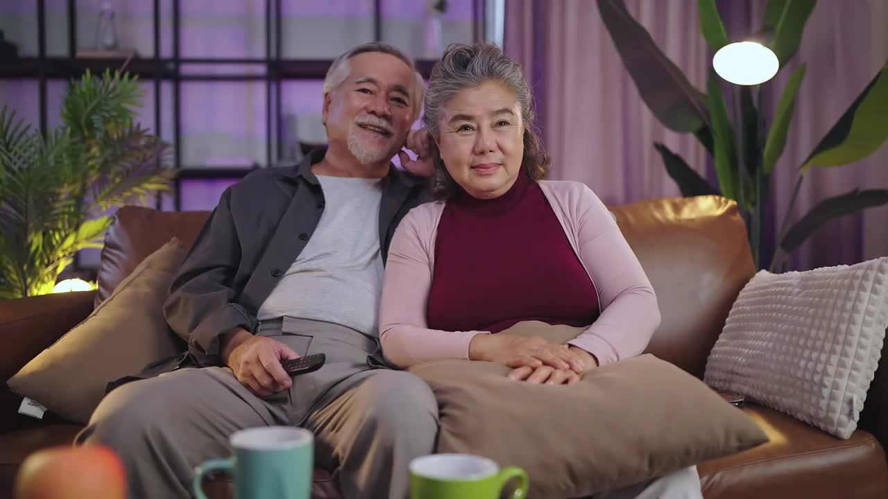 老退休的老亚洲夫妇在家里看电视，老成熟的亚洲夫妇一起在客厅的沙发沙发上欢笑胜利的体育比赛助威活动视频购买