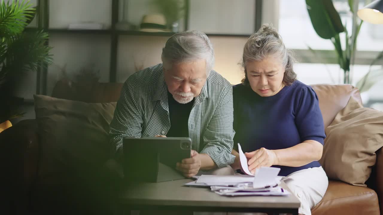 退休的亚洲老夫妇在沙发上一起检查和计算财务账单，参与财务文书工作，在客厅用电子银行笔记本电脑在线纳税视频购买
