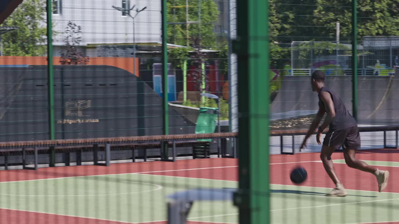 年轻的运动的非洲裔美国人打篮球，训练独自与球在室外球场，透过栅栏视频素材
