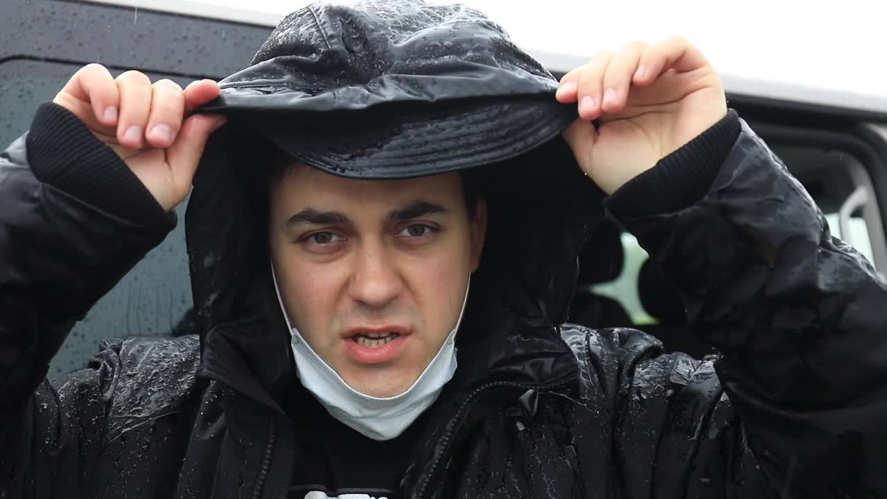 一名身穿黑色夹克的男子在大雨中戴着医用口罩，挥手致意。视频素材
