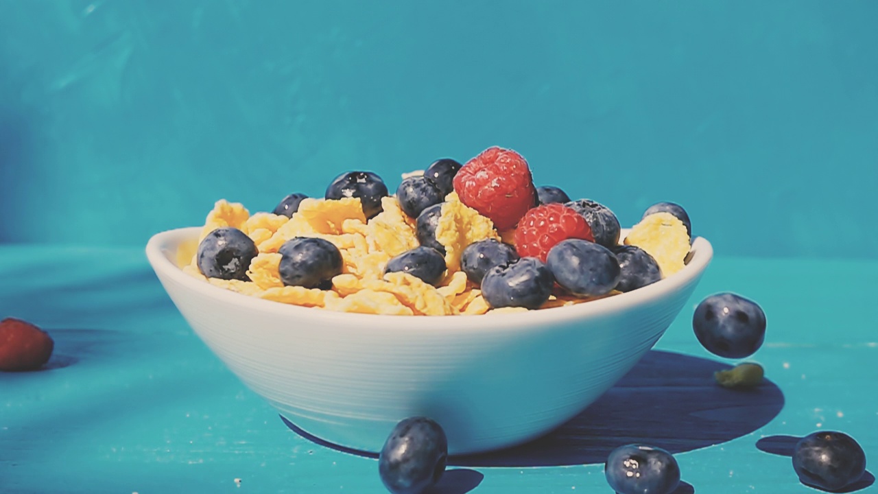 在一个阳光明媚的日子里，蓝莓和覆盆子落在一个白色的盘子里，配上黄色的玉米片，在自然的蓝色背景下放在木桌上。4k慢动作视频速度斜坡效果。视频素材
