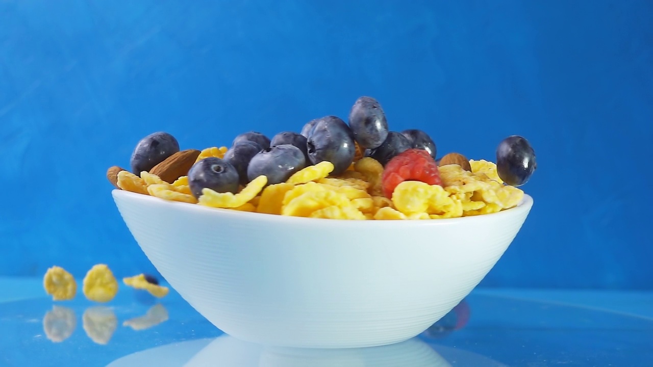 蓝莓、树莓、葡萄、杏仁和桃子的浆果落在一个旋转的白色盘子里，蓝色背景上放着黄色的玉米片。4k慢动作视频速度斜坡效果。视频素材