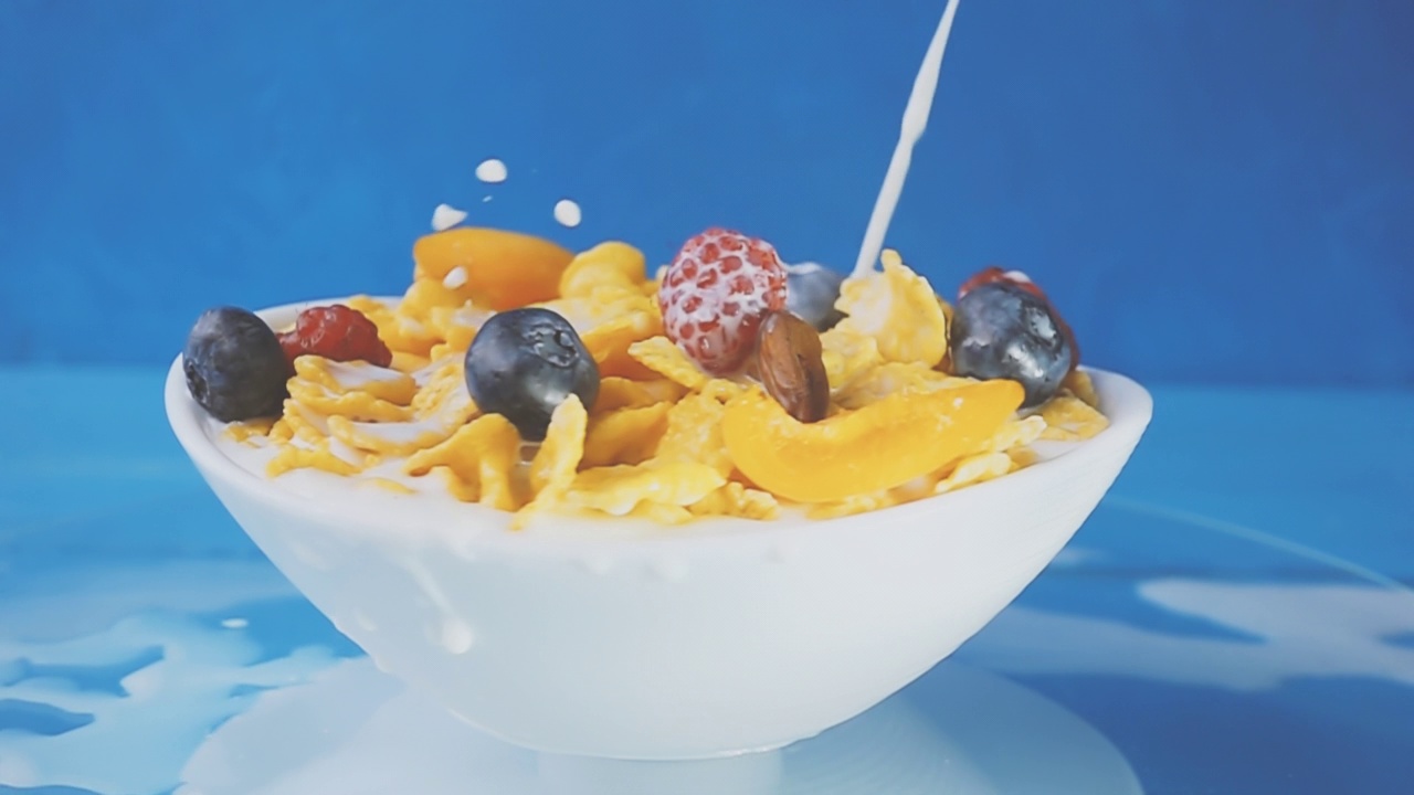 牛奶倒进黄色的玉米片里，搭配新鲜的蓝莓、覆盆子、葡萄和杏仁，放在一个浅蓝色背景的白色旋转盘里。4k原始慢动作速度斜坡视频。视频素材