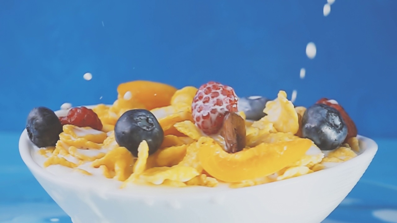 牛奶被倒进黄色的玉米片里，混合着蓝莓、覆盆子、葡萄和杏仁等新鲜浆果，放在一个蓝色背景的白色旋转盘子里。原始慢动作速度斜坡视频。视频素材