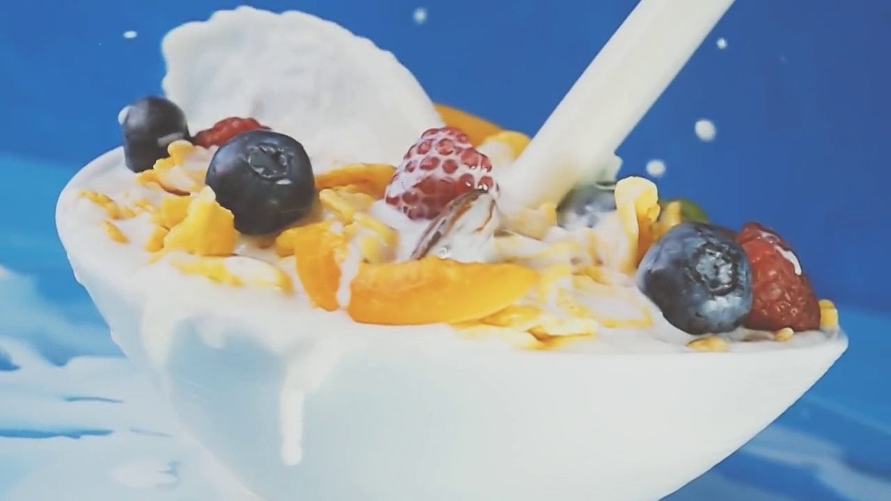 牛奶被倒进黄色的玉米片里，混合着蓝莓、覆盆子、葡萄和杏仁等新鲜浆果，放在一个蓝色背景的白色旋转盘子里。原始慢动作速度斜坡视频。视频素材