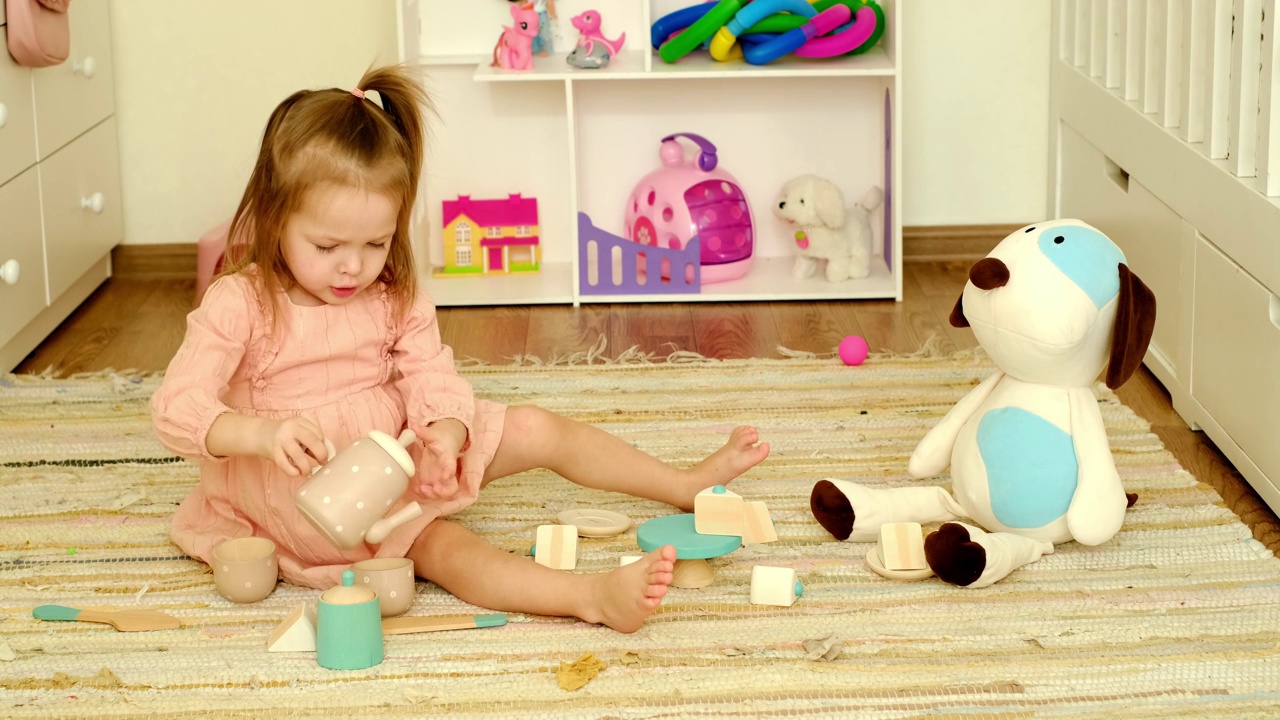 在一个茶话会上，一个穿着裙子的一岁小女孩在托儿所的地板上和一只长毛绒小狗玩耍。视频素材