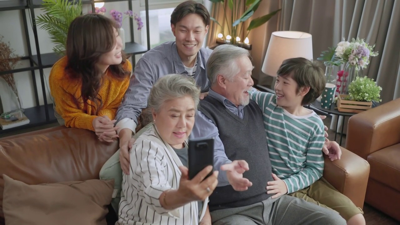 笑容满面的亚洲大家庭和孩子们坐在客厅的沙发上一起在手机上自拍视频，快乐的父母和孩子们在家里用智能手机进行有趣的视频通话视频购买