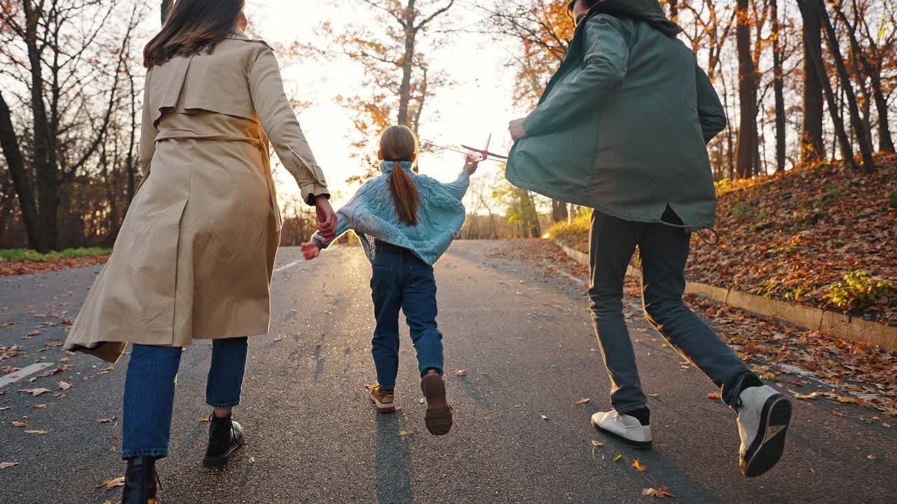 年轻的妈妈和爸爸跑着小女儿拿着红色的玩具飞机。秋天的公园。特写，慢动作。跟踪相机拍摄，后视图视频购买