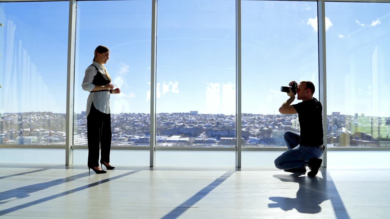 有吸引力的模特在商务西装摆相机。一名男摄影师正在拍摄一名年轻女子戴着眼镜坐在大窗户的办公室里的照片。视频下载