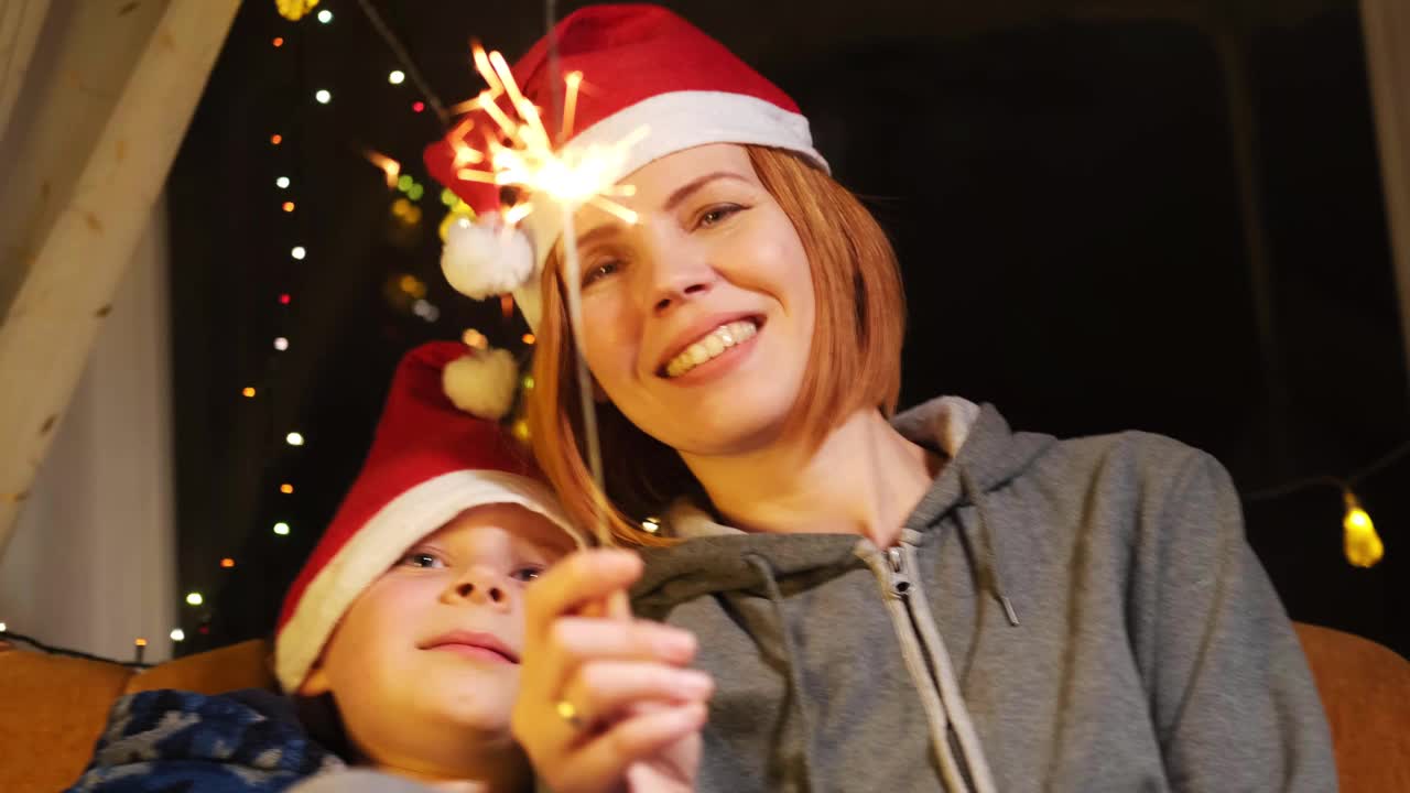 新年心情概念。妈妈和儿子戴着圣诞帽微笑着看着燃烧的烟火。视频素材