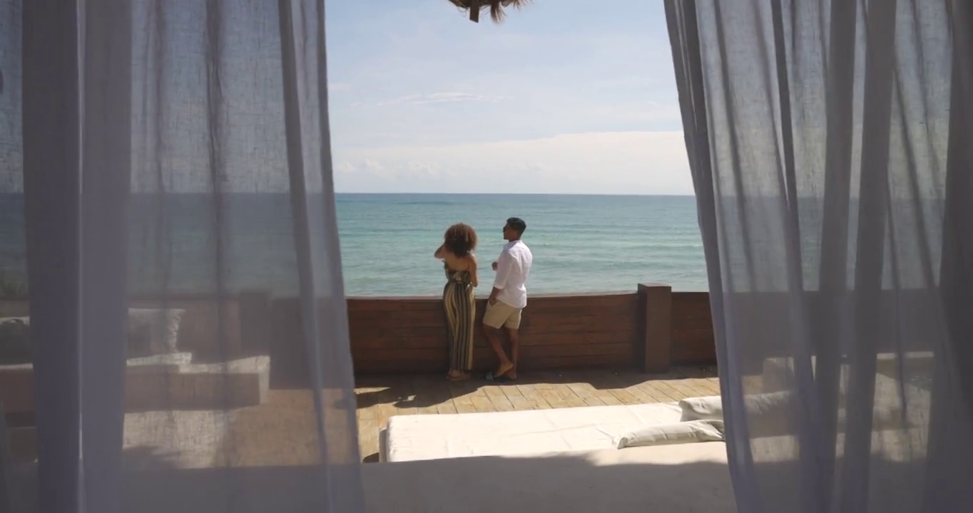 透过窗帘拍摄的一对微笑的夫妇在豪华的热带别墅甲板上享受海景视频素材