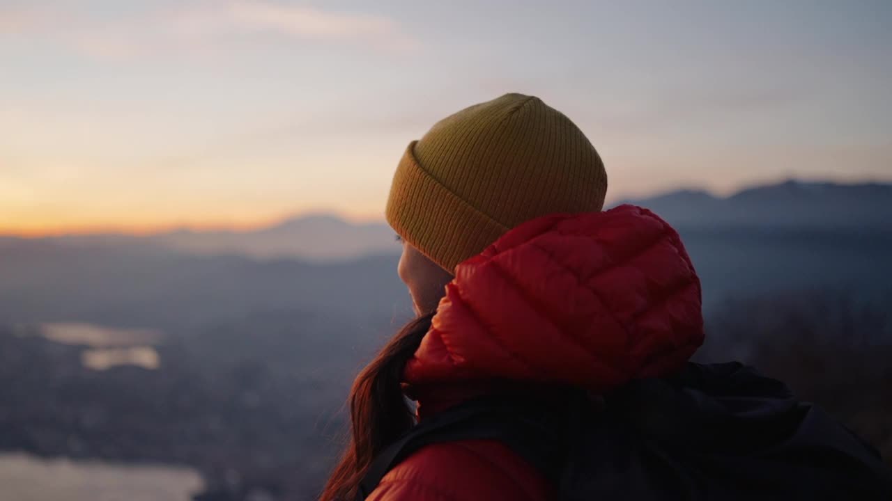 女登山站在山顶享受新鲜的空气和湖，日落和下面的城市视频下载
