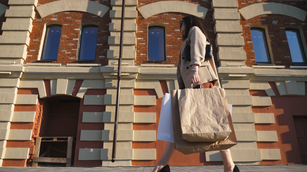 漂亮的年輕女士拿著購物袋走在人行道上買完東西。時尚的女人提著紙包走在城市的街道上。休閑和自由時間的概念。低視角慢動作視頻下載