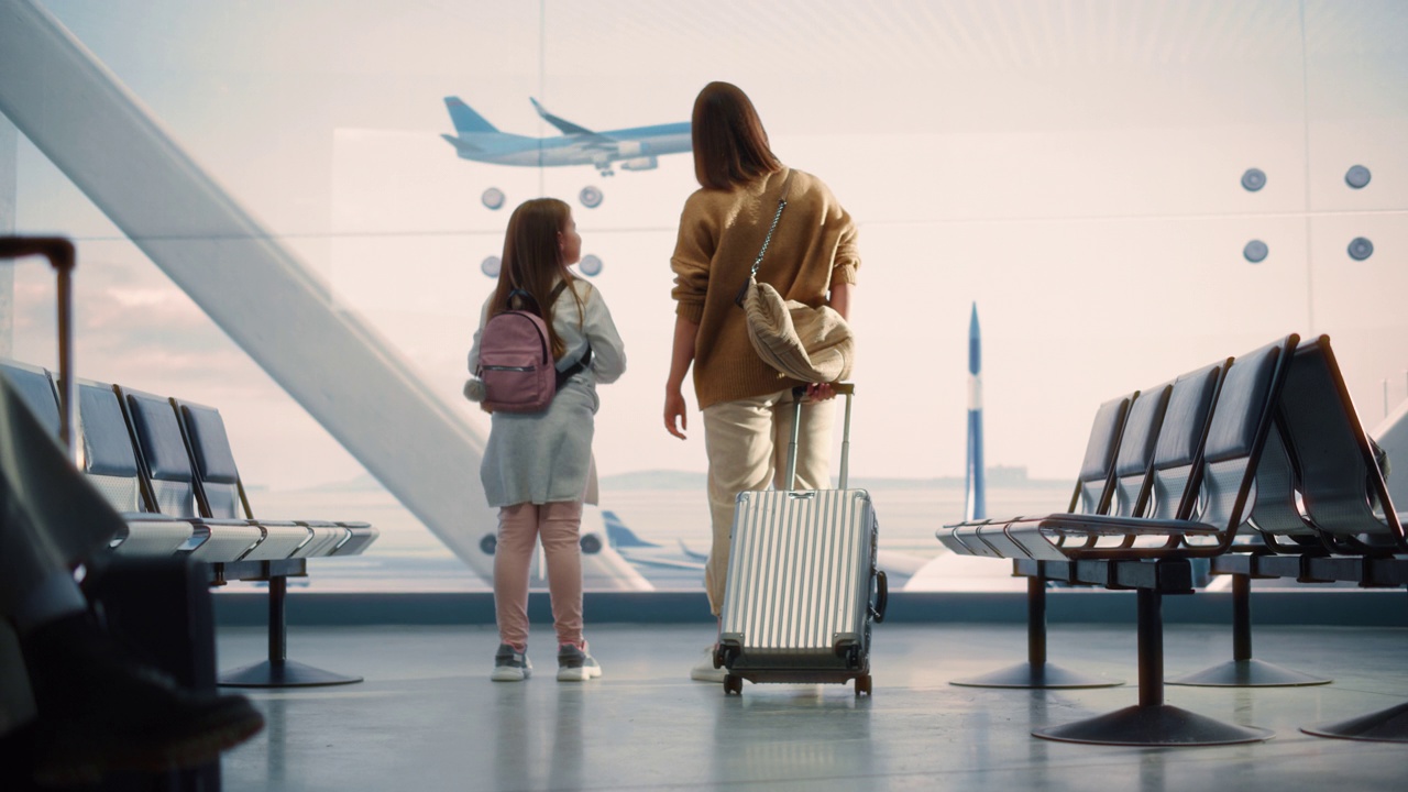 机场候机楼:美丽的母亲和可爱的小女儿在等待他们的假期航班，看着窗外到达和离开的飞机。航空枢纽登机休息室的年轻视频下载