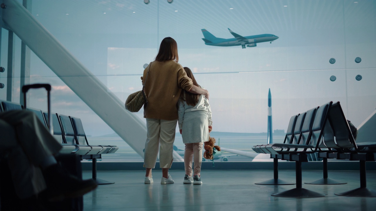 机场候机楼:美丽的母亲和可爱的小女儿在等待他们的假期航班，看着窗外到达和离开的飞机。航空枢纽登机休息室的年轻视频下载