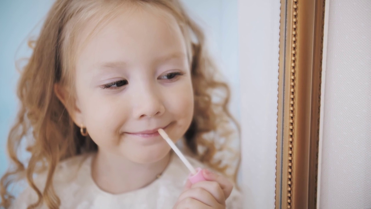 孩子画的嘴唇。肖像。可爱的卷发金发小女孩在镜子前用口红涂嘴唇视频素材