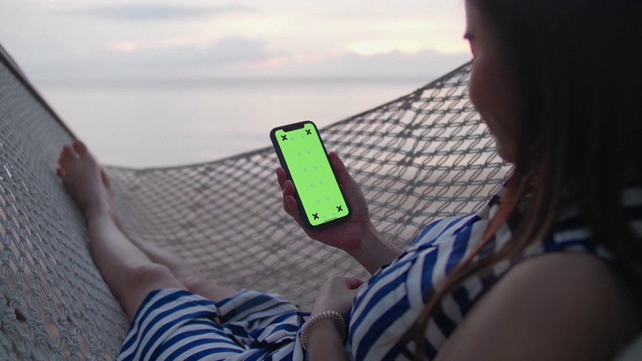 一个年轻的女孩躺在沙滩上的吊床上使用智能手机视频下载