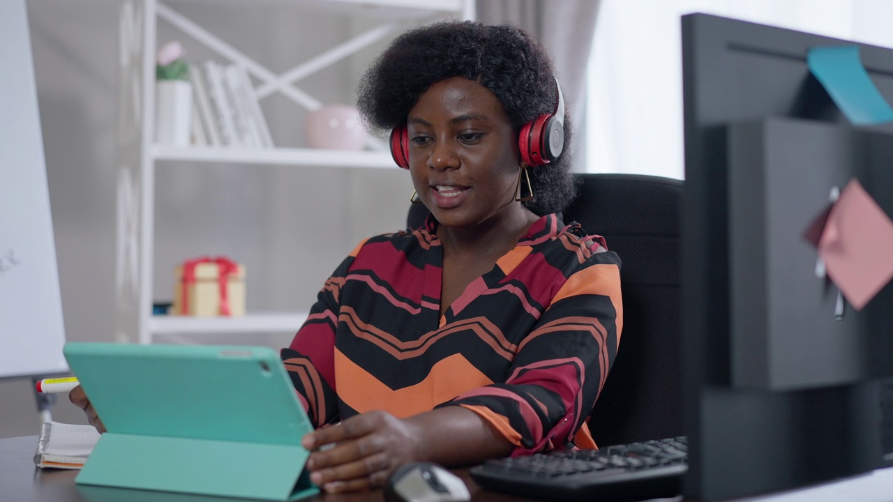 积极自信的非洲裔美国妇女戴着耳机，坐在家庭办公室里用平板电脑视频聊天。成功的年轻自由职业者的肖像以慢动作微笑交谈。远程工作。视频素材