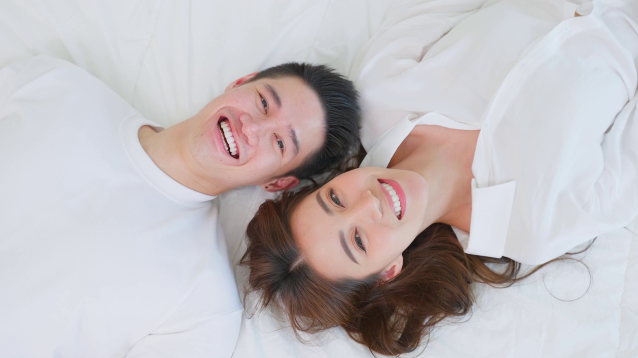 亞洲新婚夫婦躺在床上看著相機的肖像。漂亮漂亮的年輕男女穿著睡衣在家里的臥室里享受清晨的活動。關系的概念視頻購買