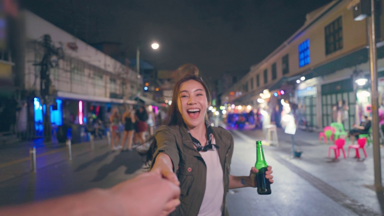 亚洲女游客的肖像，牵着男友的手，走在街上。在城市旅游的年轻男女花时间在一起度假，在漆黑的夜晚在路上喝一瓶啤酒。视频素材