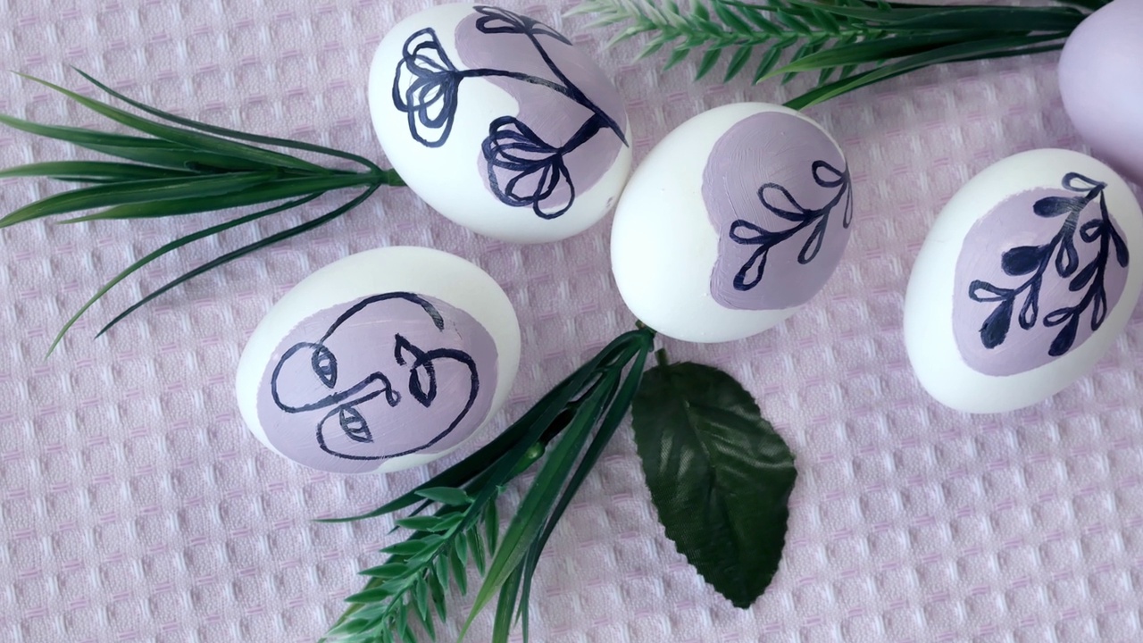复活节的概念。在一条线上画有一张脸的白色鸡蛋，紫色上画有植物。绿叶，厨房毛巾。照相机沿着构图平移视频下载