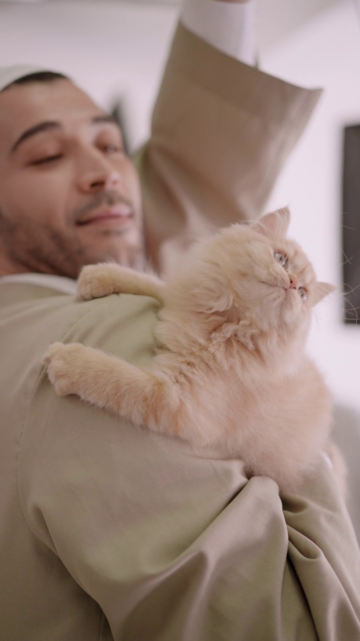 侧视图:穿着宗教服装的英俊穆斯林男子肩膀上的一只波斯小猫，像孩子一样抱着小猫的胸膛，用猫玩具玩耍视频下载