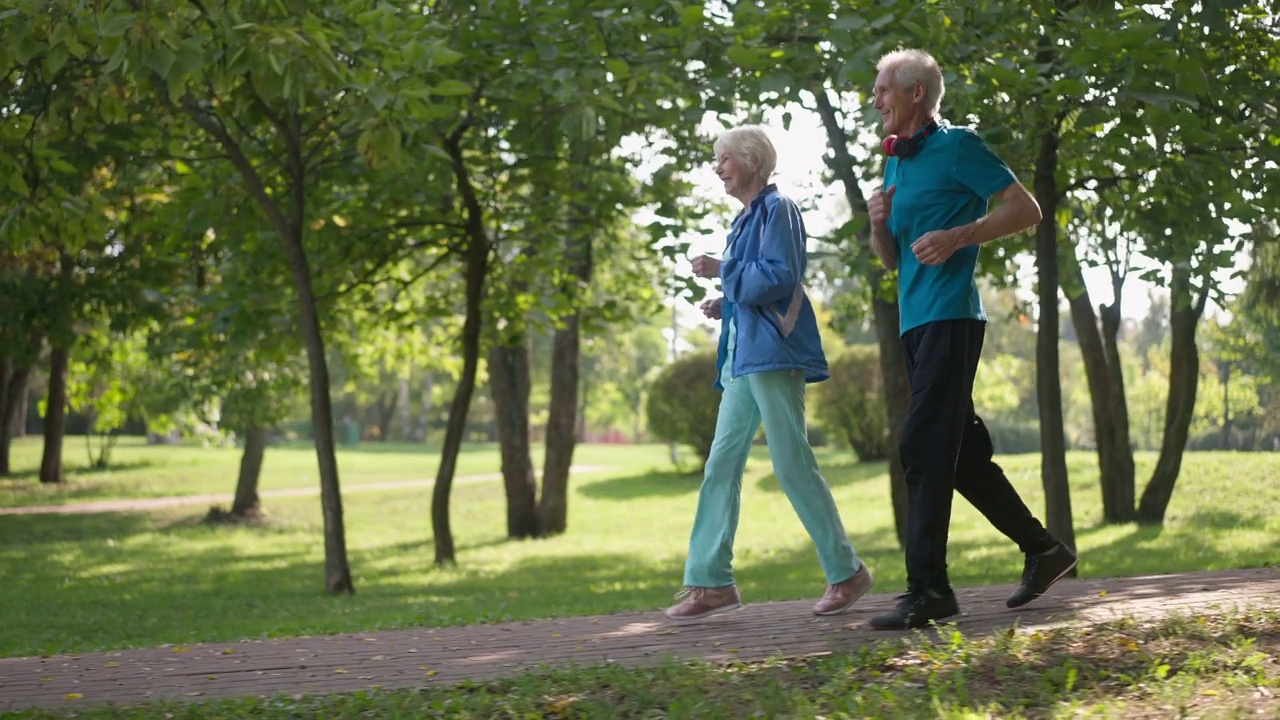 侧视图快乐的老年夫妇跑步慢动作在阳光下户外微笑。广角镜头，自信的高加索男女在阳光明媚的春天夏天的公园慢跑。体育和爱情。视频下载