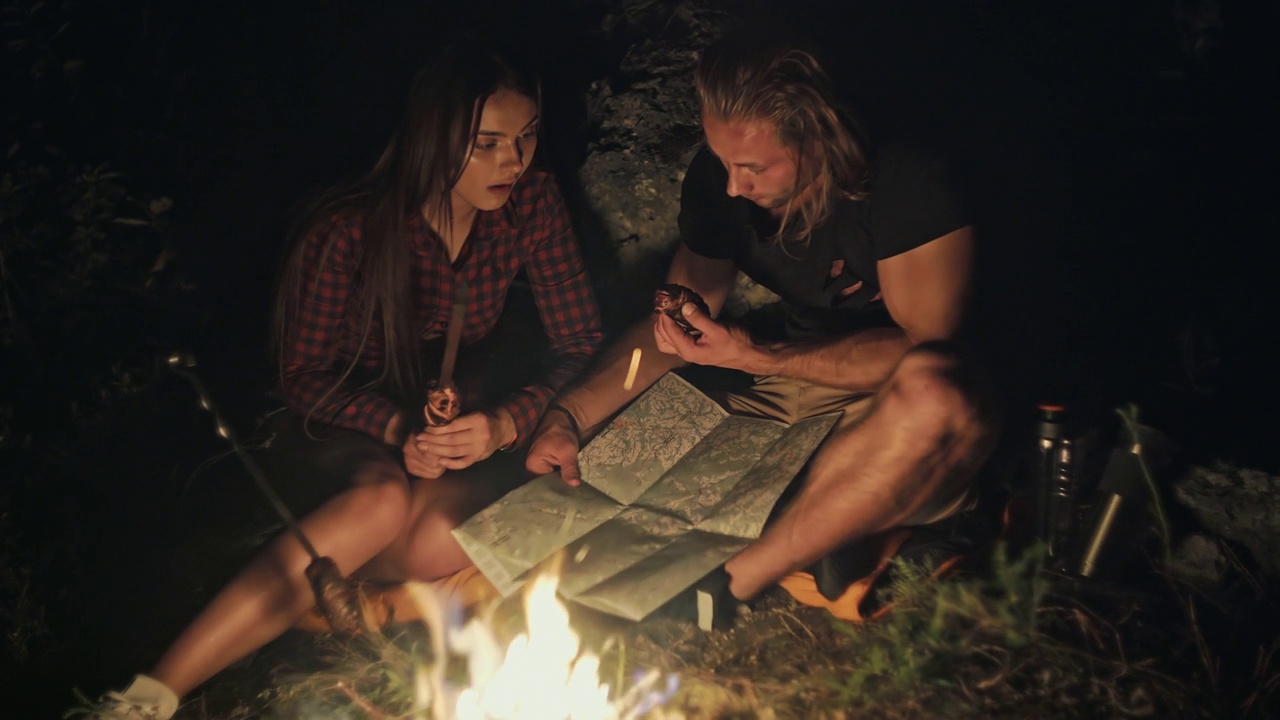 漂亮的年轻夫妇在森林里的篝火旁休息，晚上仔细地寻找地图和晚餐视频下载