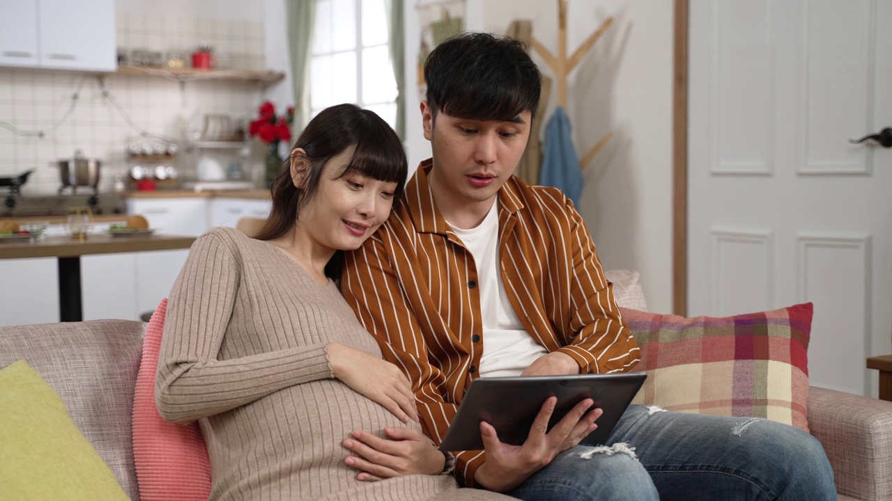 幸福的亚洲怀孕夫妇在家里客厅的沙发上用电子平板一起网购婴儿用品视频下载