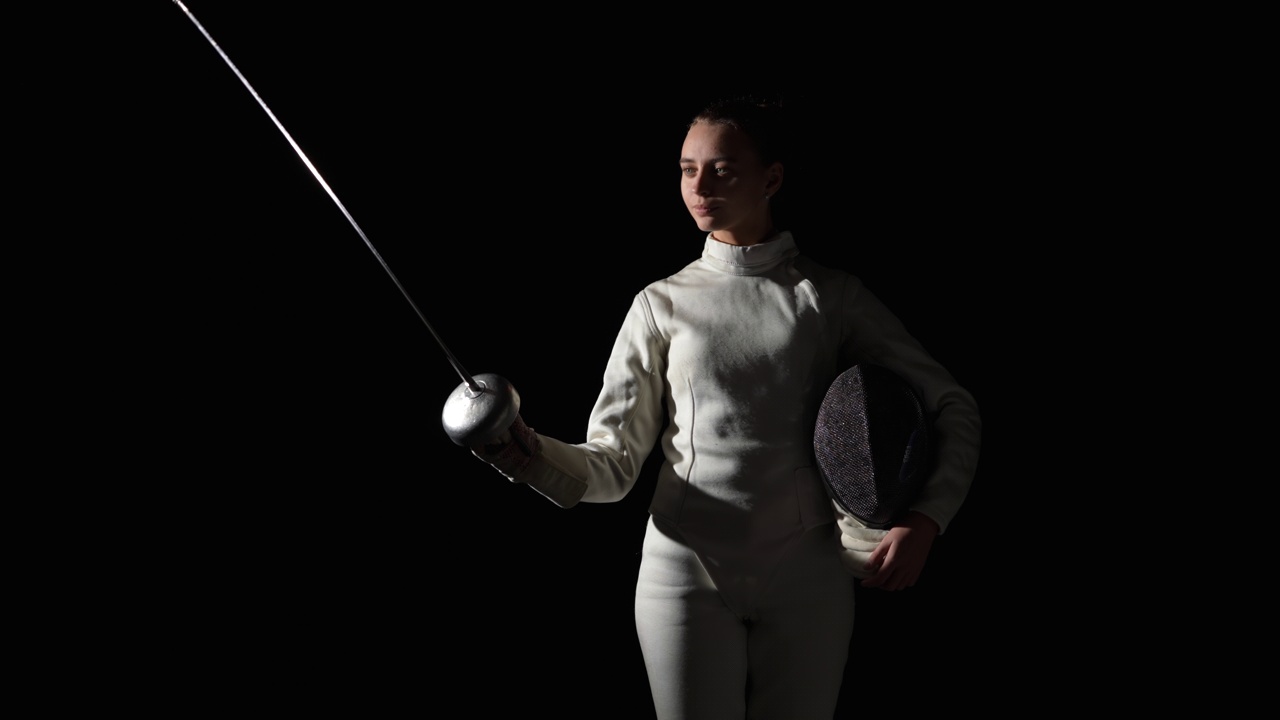 一名年轻女子击剑手在问候时用剑敬礼的肖像。一名身穿白色制服的运动员在黑暗的演播室里摆姿势，背景是黑色的，灯光是舞台上的。缓慢的运动。近距离视频下载