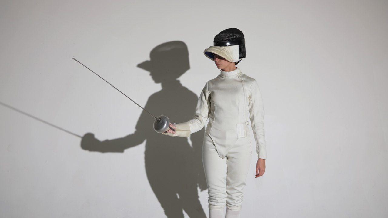 一名年轻女子击剑手的肖像，戴上头盔，进入战斗位置。一名运动员在白色背景和阴影的黑暗工作室里摆姿势。缓慢的运动。近距离视频下载
