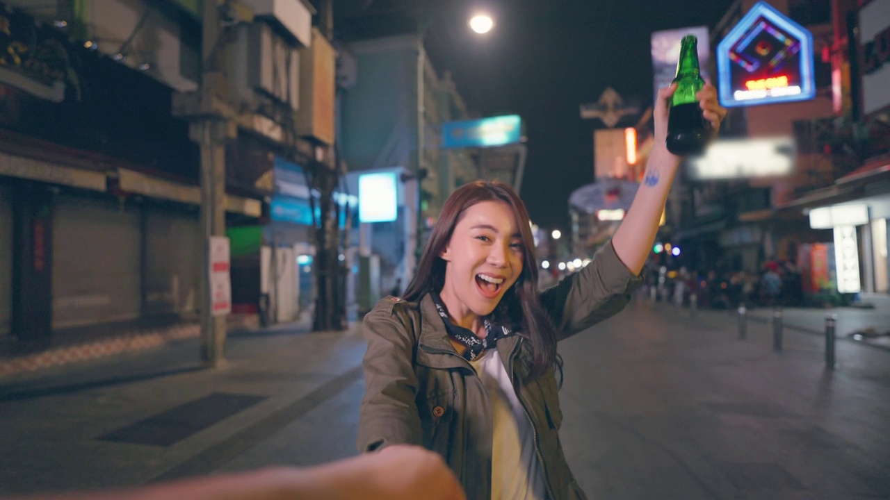 亚洲女游客的肖像，牵着男友的手，走在街上。在城市旅游的年轻男女花时间在一起度假，在漆黑的夜晚在路上喝一瓶啤酒。视频下载