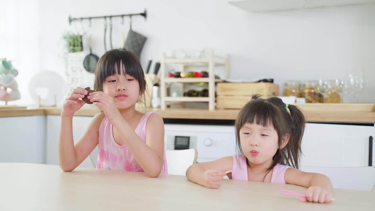 亚洲两个可爱的小女孩姐妹喜欢吃快餐，薯片。饥肠辘辘的学前班小女孩坐在厨房的桌子上，用手把零食放进嘴里。美味又不健康的儿童食品理念。视频购买
