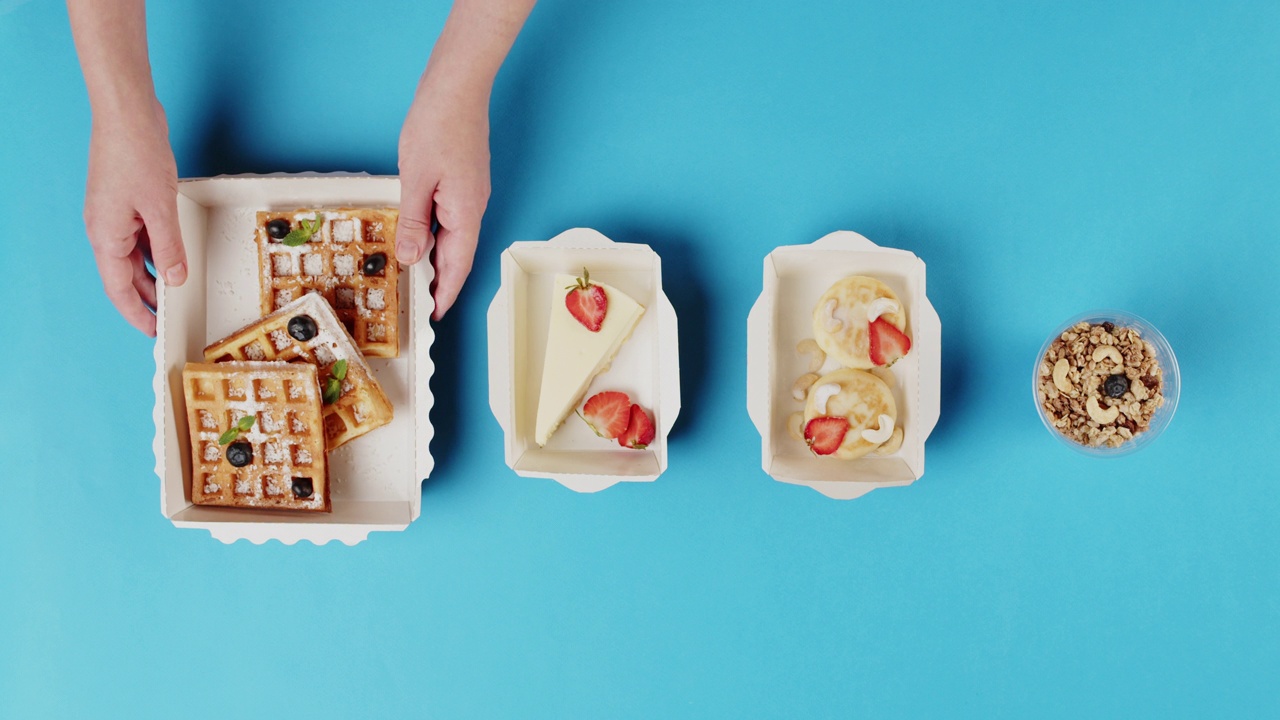 食品配送俯视图，在蓝色背景的一次性容器中带走甜点。带芝士蛋糕的午餐盒，比利时华夫饼。餐饮服务糖果。视频购买