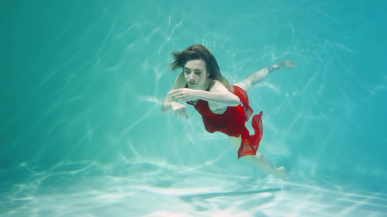 年轻女子在红色雪纺服装跳舞水下蓝色的水柱与眩光。女舞者在海水中跳舞，浮到水面呼吸空气。慢动作特写视频下载