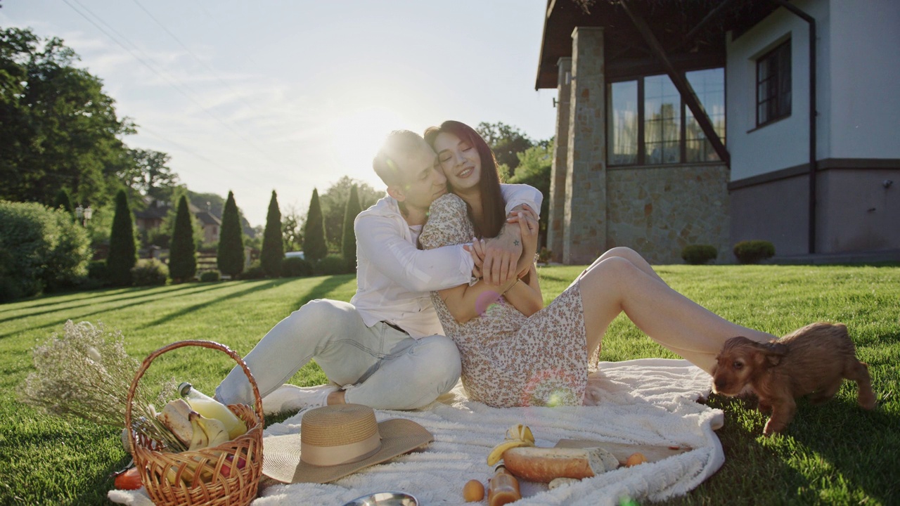 浪漫的野餐男孩和女孩坐在漂亮房子附近的草坪上，快乐地拥抱着小狗玩耍视频下载