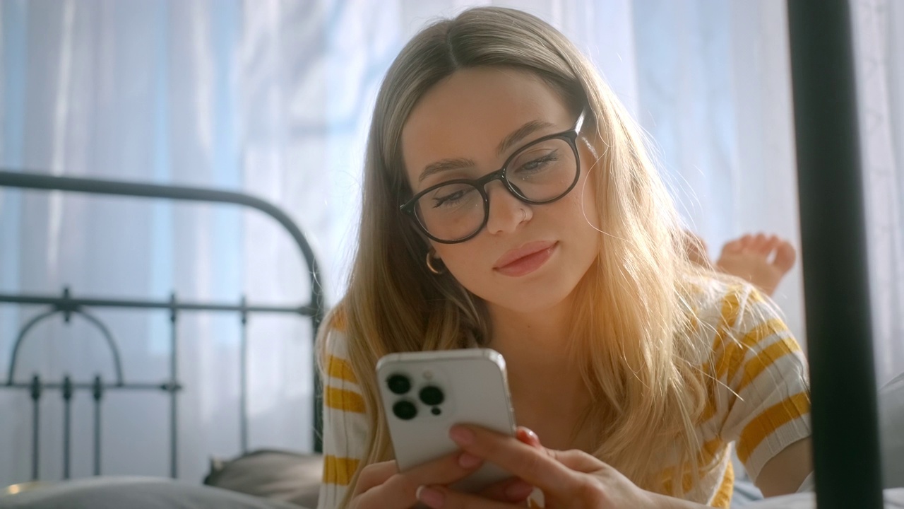 快乐微笑的年轻迷人的白人妇女戴着眼镜使用智能手机，在阳光明媚的日子里躺在床上。视频下载