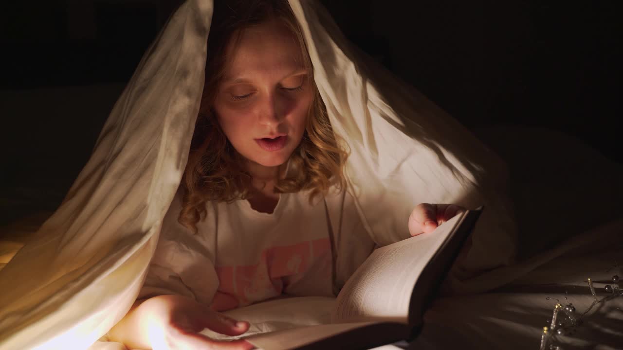 女人在黑暗中盖着毯子看书，并用手电筒照亮书页。她对自己读到的东西感到惊讶，微笑着说。阅读，童话故事，学习一些东西视频下载