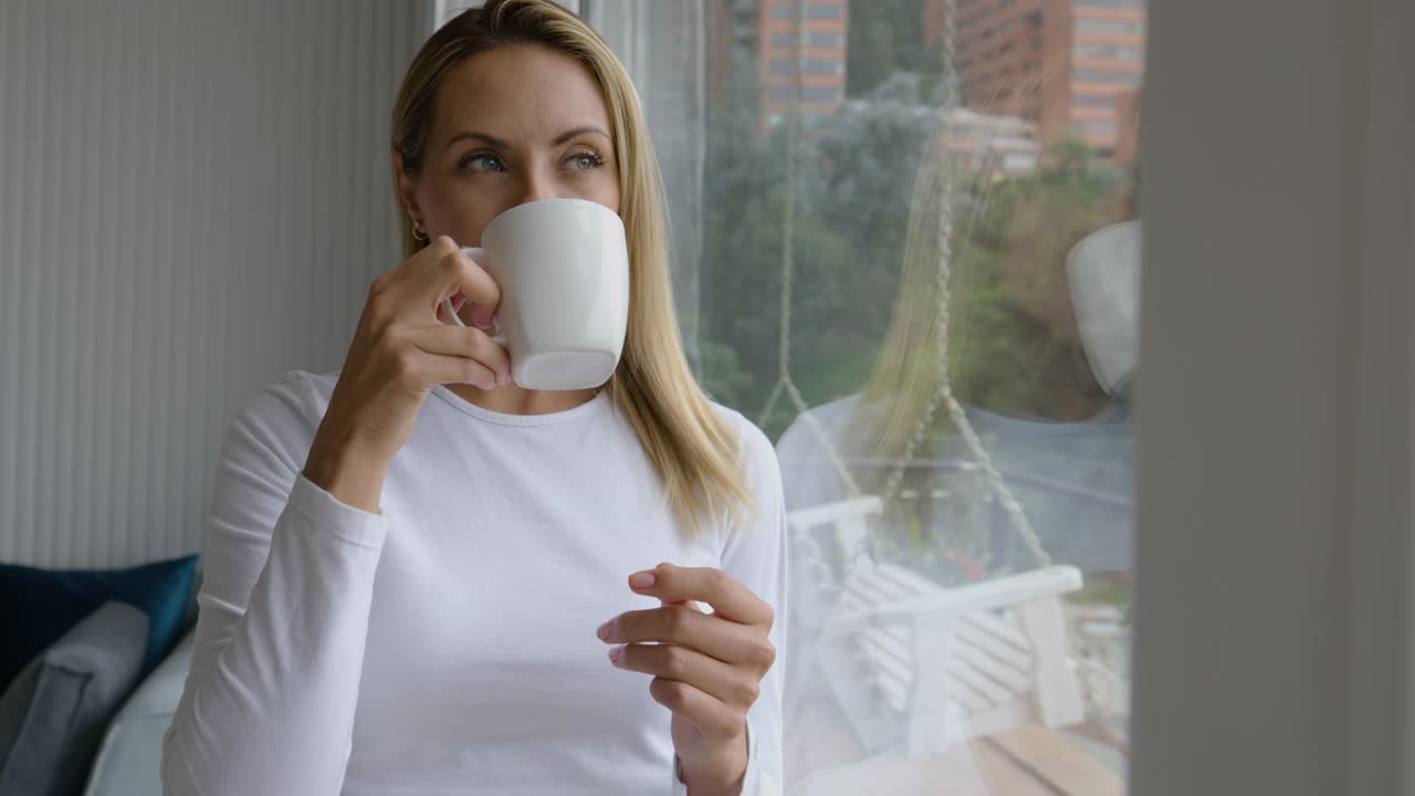 美丽的女人在家里穿着睡衣，一边喝着咖啡，一边看着窗外的风景发呆视频素材