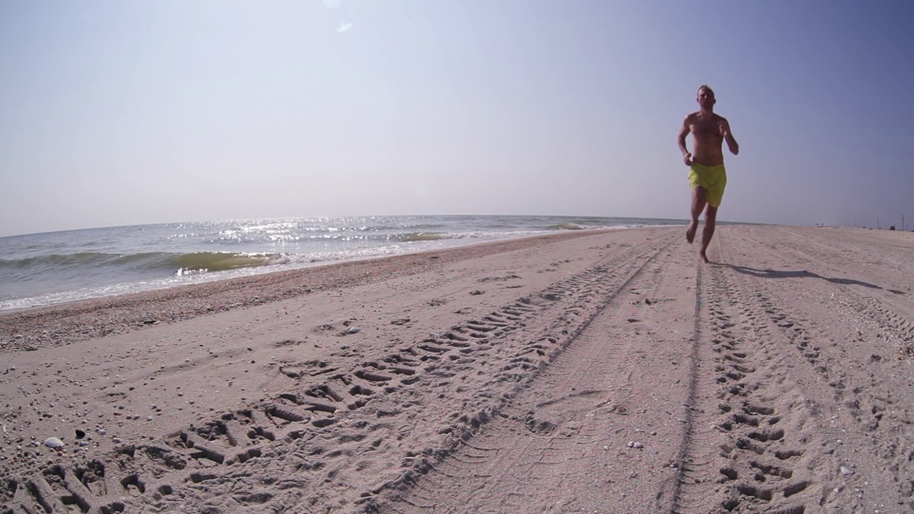 阳光明媚的一天，一名男子走近摄像机，在沙滩上奔跑。海滨度假，跑步，自然理念。视频素材
