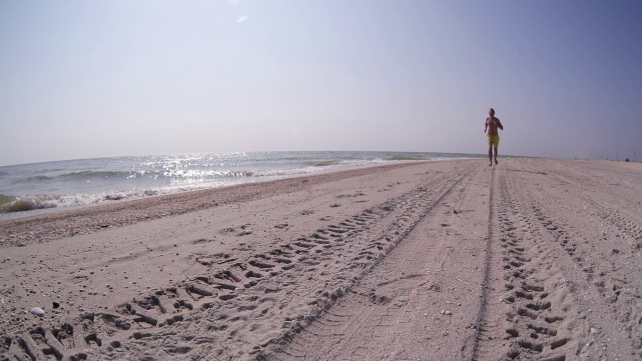 阳光明媚的一天，一名男子跑过沙滩，走近摄像机。海滨度假，跑步，自然概念镜头。视频素材