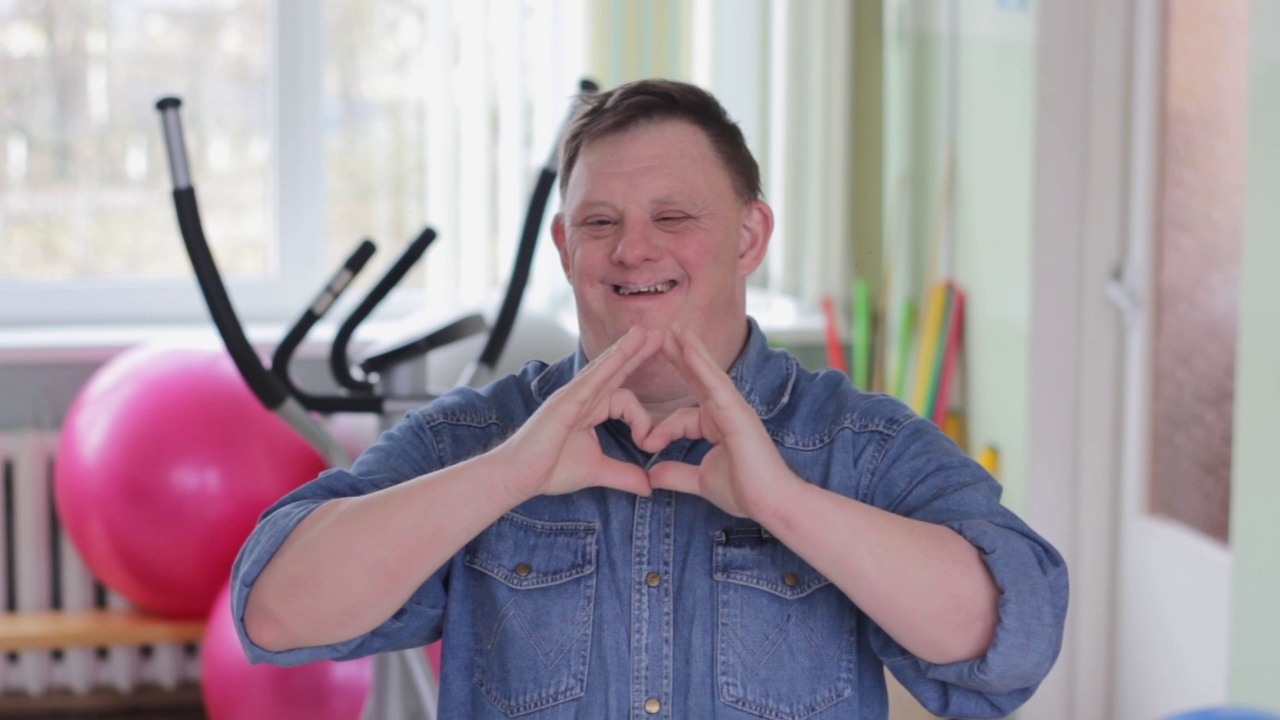 一位患有唐氏综合症的残疾人对着镜头微笑，用手画出了一个心形。视频下载